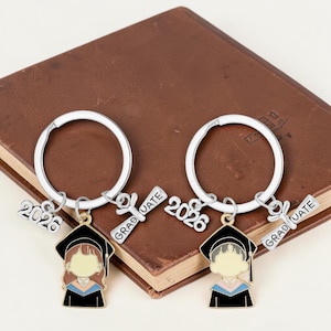 May include: Two graduation-themed keychains with silver rings. Each keychain features a miniature graduate in a black cap and gown, a diploma charm with "GRADUATE" and a "2026" charm. The keychains are resting on a brown book.