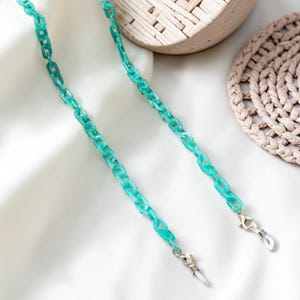 Puede incluir: Una cadena turquesa de acrílico para gafas. La cadena está compuesta por eslabones rectangulares y presenta cierres plateados en cada extremo para sujetar las gafas. La cadena se muestra sobre una superficie blanca.