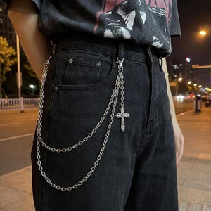 May include: Black denim jeans featuring a silver-toned metal wallet chain attached to the belt loop. The chain consists of two draped strands and a decorative cross pendant with intricate detailing. The jeans have a classic five-pocket design.