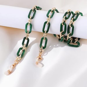 Op de afbeelding: Een groene en gouden brillenketting. De ketting heeft rechthoekige groene schakels die zijn verbonden door goudkleurige decoratieve elementen. De ketting is ontworpen om een bril vast te houden, met witte rubberen lussen aan elk uiteinde om aan de bril te bevestigen.