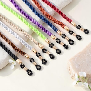 Op de afbeelding: Een verzameling kleurrijke brillenkoorden in verschillende tinten, waaronder zwart, bruin, wit, goud, groen, paars, blauw, oranje en rood. Elke ketting heeft een gedraaid touwontwerp en goudkleurige connectoren met zwarte rubberen lussen.