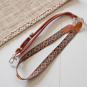 Könnte beinhalten: Ein brauner und gemusterter Taschenriemen mit einem passenden braunen Kordelriemen. Der gemusterte Riemen weist ein sich wiederholendes geometrisches Design in Braun-, Orange- und Weißtönen auf. Silberfarbene Metallverschlüsse und Akzente sind sichtbar.