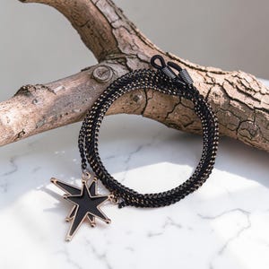 Könnte beinhalten: Schwarze und goldene Brillenketten mit Sternanhänger. Die Kette besteht aus einem gewebten schwarzen und goldenen Material. Der Sternanhänger ist schwarz mit goldenem Rand. Die Kette ist um ein Stück Holz geschlungen.