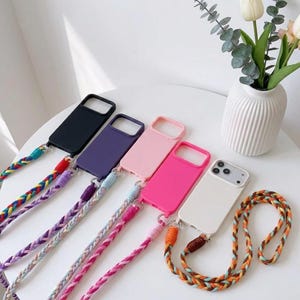 May include: Five rectangular smartphone cases in black, purple, light pink, hot pink, and white, each featuring a thick, braided cord strap. The straps display various color combinations including rainbow, purple, pink, and earth tones with metal rings.