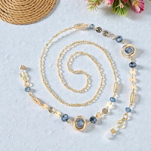 May include: Gold-coloured glasses chain with blue and gold beads. The chain has a delicate design with small, round gold accents and larger, oval blue beads. The chain is designed to hold spectacles.