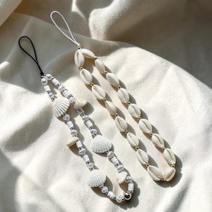 May include: Two phone charms featuring white cowrie shells and beads. One charm has a black cord and includes white clam-shaped shells, small white pearls, and cylindrical beads. The other charm has a white cord and a single strand of cowrie shells.