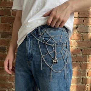 May include: A silver-toned metal body chain featuring a spider web design draped over blue denim jeans. The accessory consists of interconnected thin chains forming a web pattern with a small spider charm attached to the lower centre section.