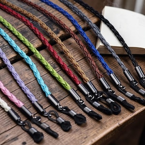 Puede incluir: Una colección de cordones trenzados para gafas en varios colores: rosa, morado, azul claro, verde, rojo, marrón, azul, gris y negro. Cada cordón tiene conectores metálicos plateados y bucles de goma negros para sujetar las gafas. Dispuestos sobre una superficie de madera.