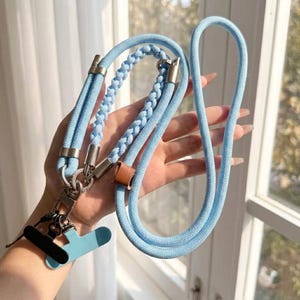 May include: A light blue braided rope lanyard featuring silver-toned metal hardware, a small brown leather accent, and two attached clips in black and light blue. The cord is constructed from a smooth, woven material with a consistent texture throughout.