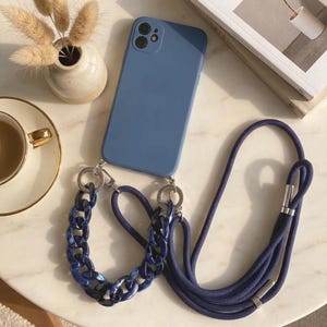 May include: A blue smartphone case with a dual-camera cutout, featuring a detachable strap system. The strap consists of a thick, dark blue rope cord and a chunky, translucent navy blue acrylic chain link section, both connected by silver-toned metal rings.