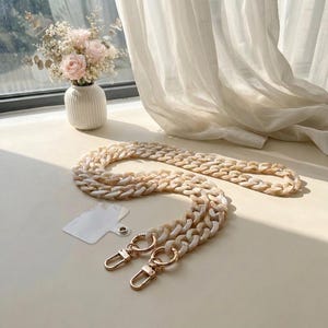 May include: A phone strap featuring thick, interlocking beige and white marbled acrylic chain links. The strap includes two gold-toned metal lobster claw clasps attached to circular rings and a clear, rectangular plastic phone anchor tab.