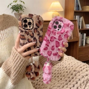 Può includere: Due cover per telefono soffici con aperture per la fotocamera. Una cover presenta un motivo leopardato marrone e nero, mentre l'altra è rosa acceso con un motivo leopardato abbinato. Entrambe includono un ciondolo composto da tre piccoli pon-pon.