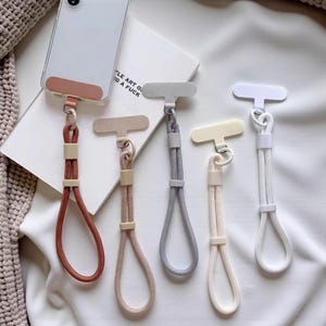 Op de afbeelding: Vijf telefoonlanyards in roest-, roze-, grijze, crème- en witte tinten. Elk heeft een handlus en een T-vormige lip om in een telefoonhoesje te steken. De lanyards zijn gerangschikt op een witte ondergrond. De tekst op het boek luidt "LE ART OF G A FUCK".