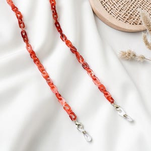 Op de afbeelding: Rode en roze brillenketting met zilverkleurige sluitingen. De ketting is gemaakt van rechthoekige schakels en is ontworpen om een bril vast te houden. De achtergrond is een witte stof.
