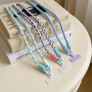 May include: Three braided phone lanyards featuring pastel colour palettes. Each strap is constructed from woven fabric cords in shades of light blue, white, pink, and lavender, finished with a solid-coloured plastic attachment tab at the base.