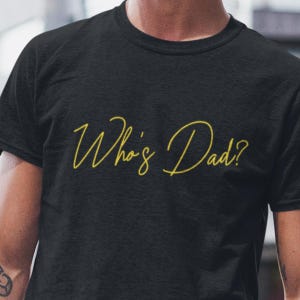 Op de afbeelding: Zwart T-shirt met de tekst "Who's Dad?" in gele sierlijke letters. Het shirt heeft een ronde hals en lijkt gemaakt van een zacht materiaal. De tekst is gecentreerd op de voorkant van het shirt.