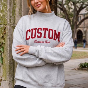 May include: A light gray long-sleeve sweatshirt with a mock neck collar. The front features the word CUSTOM in large, bold, dark red block letters, with the words Custom Text printed underneath in a smaller, cursive, dark red font.