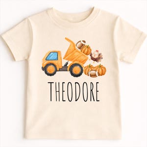 May include: A cream-colored short-sleeve t-shirt featuring a watercolor graphic of an orange dump truck carrying a football and a pumpkin. Beside the truck are two more pumpkins, a football, and a turkey. The name THEODORE is printed in black.