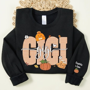 May include: A black long-sleeve sweatshirt featuring the word GIGI in large orange and white gingham letters. The design includes a small orange beanie, a pumpkin, and boots with faces. The sleeve displays the names Sophia and Liam with a pumpkin.