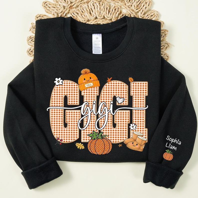 May include: A black long-sleeve sweatshirt featuring the word GIGI in large orange and white gingham letters. The design includes a small orange beanie, a pumpkin, and boots with faces. The sleeve displays the names Sophia and Liam with a pumpkin.