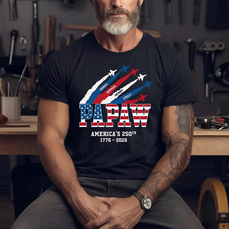 May include: Navy blue t-shirt with a patriotic design. The shirt features the word "PAPAW" in red, white, and blue, with the text "AMERICA'S 250TH 1776-2026" below. Above the word "PAPAW" are jets with names.
