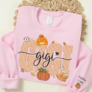 May include: A light pink crewneck sweatshirt featuring the word GIGI in large orange and white gingham letters. The design includes a small orange beanie, a pumpkin, and boots. The sleeve displays the names Sophia and Liam with a small pumpkin.