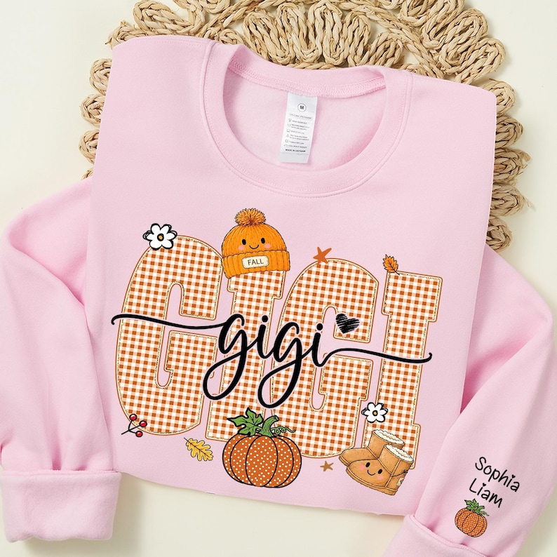 May include: A light pink crewneck sweatshirt featuring the word GIGI in large orange and white gingham letters. The design includes a small orange beanie, a pumpkin, and boots. The sleeve displays the names Sophia and Liam with a small pumpkin.