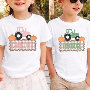 May include: Two white t-shirts featuring a graphic print of a tractor, two orange pumpkins, and a brown gingham rectangle. One shirt displays the name Charlotte in pink, while the other displays the name Theodore in green, both in a sans-serif font.