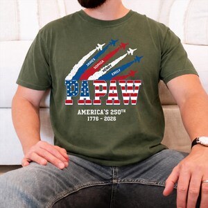 May include: Olive green t-shirt with a patriotic design. The shirt features the word "PAPAW" in red, white, and blue, with the text "AMERICA'S 250TH 1776-2026" below. Above the word "PAPAW" are jets with the names James, Sophia, Henry, and Emily.