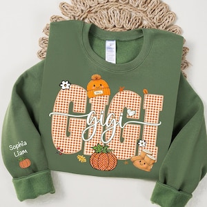 May include: An olive green crewneck sweatshirt featuring a large orange and white gingham graphic with the text "GIGI" in a cursive font. The design includes a smiling orange beanie, a pumpkin, and boots. The sleeve displays the names "Sophia" and "Liam" with a small pumpkin icon.