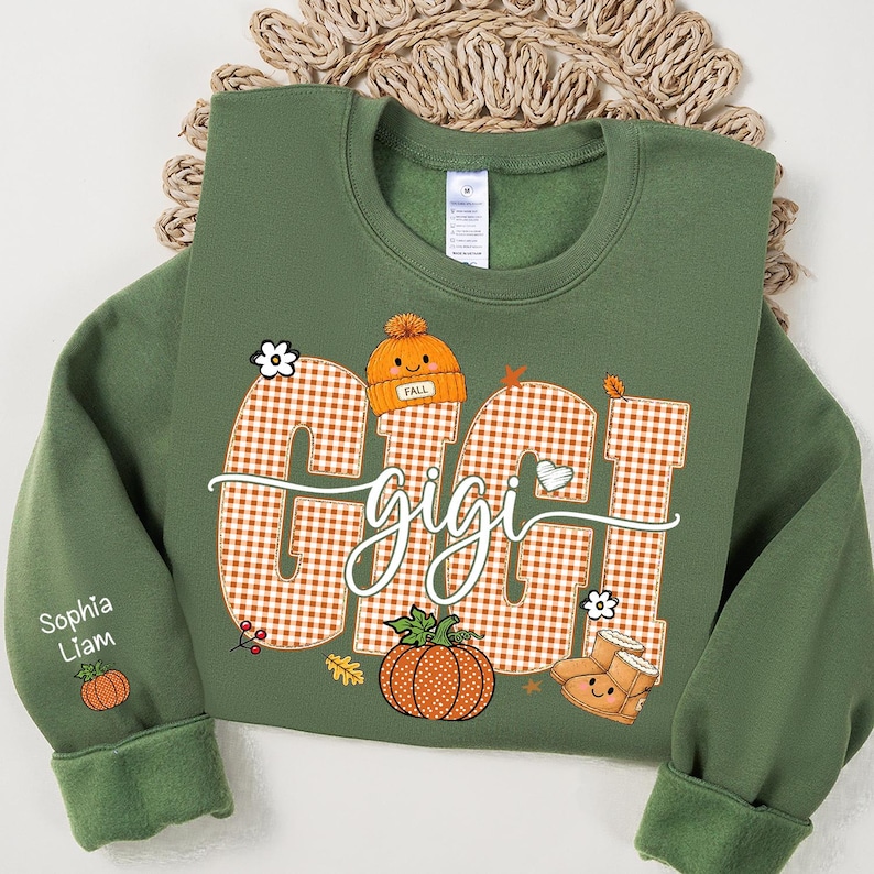 May include: An olive green crewneck sweatshirt featuring a large orange and white gingham graphic with the text "GIGI" in a cursive font. The design includes a smiling orange beanie, a pumpkin, and boots. The sleeve displays the names "Sophia" and "Liam" with a small pumpkin icon.