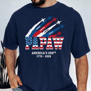 May include: Navy blue t-shirt with a patriotic design. The word "PAPAW" is in red and white stripes with a blue star-filled font. Above, jets leave trails with names. Below, text reads "America's 250th 1776-2026".