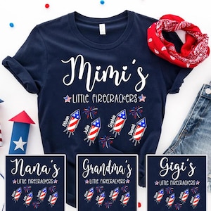 May include: A navy blue short-sleeved t-shirt featuring the white text Mimi's Little Firecrackers, accompanied by four red, white, and blue firecracker graphics labeled with the names Ethan, Julia, Sophia, and Daniel, plus fireworks.