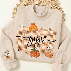 May include: A beige crewneck sweatshirt featuring a large graphic with the word GIGI in orange and white gingham block letters. The design includes a smiling orange beanie, a pumpkin, and boots. The sleeve displays the names Sophia and Liam.