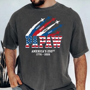 May include: Dark gray t-shirt with a patriotic design. The shirt features the word "PAPAW" in red and blue, with the American flag incorporated into the letters. Above the word are jets with white, red, and blue vapor trails, and names. The text "AMERICA'S 250TH 1776-2026" is below.