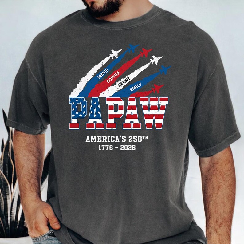 May include: Dark gray t-shirt with a patriotic design. The shirt features the word "PAPAW" in red and blue, with the American flag incorporated into the letters. Above the word are jets with white, red, and blue vapor trails, and names. The text "AMERICA'S 250TH 1776-2026" is below.