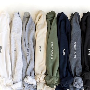 May include: A collection of eight long-sleeve crewneck sweatshirts arranged side-by-side in a row. The colors from left to right are labeled as White, Ash, Sport Grey, Sand, Military Green, Navy, Dark Heather, and Black, each featuring a ribbed collar.