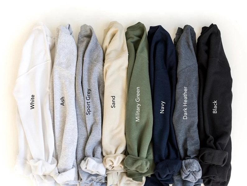 May include: A collection of eight long-sleeve crewneck sweatshirts arranged side-by-side in a row. The colors from left to right are labeled as White, Ash, Sport Grey, Sand, Military Green, Navy, Dark Heather, and Black, each featuring a ribbed collar.