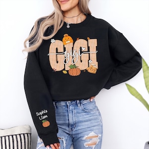May include: A black crewneck sweatshirt featuring a large graphic on the chest with the word GIGI in orange and white gingham letters. The design includes an orange beanie, a pumpkin, and autumn leaves. The sleeve displays the names Sophia and Liam.