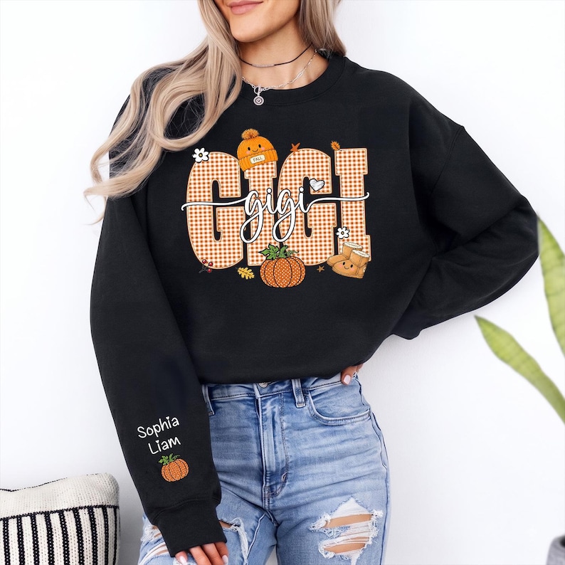 May include: A black crewneck sweatshirt featuring a large graphic on the chest with the word GIGI in orange and white gingham letters. The design includes an orange beanie, a pumpkin, and autumn leaves. The sleeve displays the names Sophia and Liam.