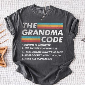 May include: A charcoal gray short-sleeve t-shirt featuring the text THE GRANDMA CODE in white, accompanied by five numbered rules including Bedtime is whenever and Hugs are mandatory, with horizontal teal, yellow, and red stripes across the chest.