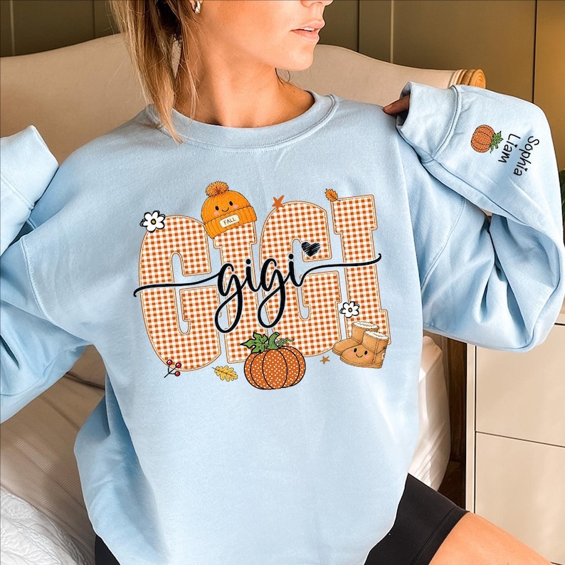 May include: A light blue crewneck sweatshirt featuring the word GIGI in large orange and white checkered letters. The design includes a pumpkin, a beanie, and boots. The sleeve is personalized with the names Sophia and Liam next to a pumpkin.
