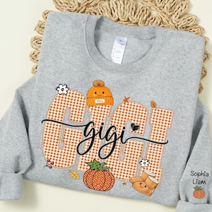 May include: A light gray crewneck sweatshirt featuring the word GIGI in large orange and white gingham letters. The design includes an orange beanie, a pumpkin, and boots with faces. The sleeve displays the names Sophia and Liam with a small pumpkin.