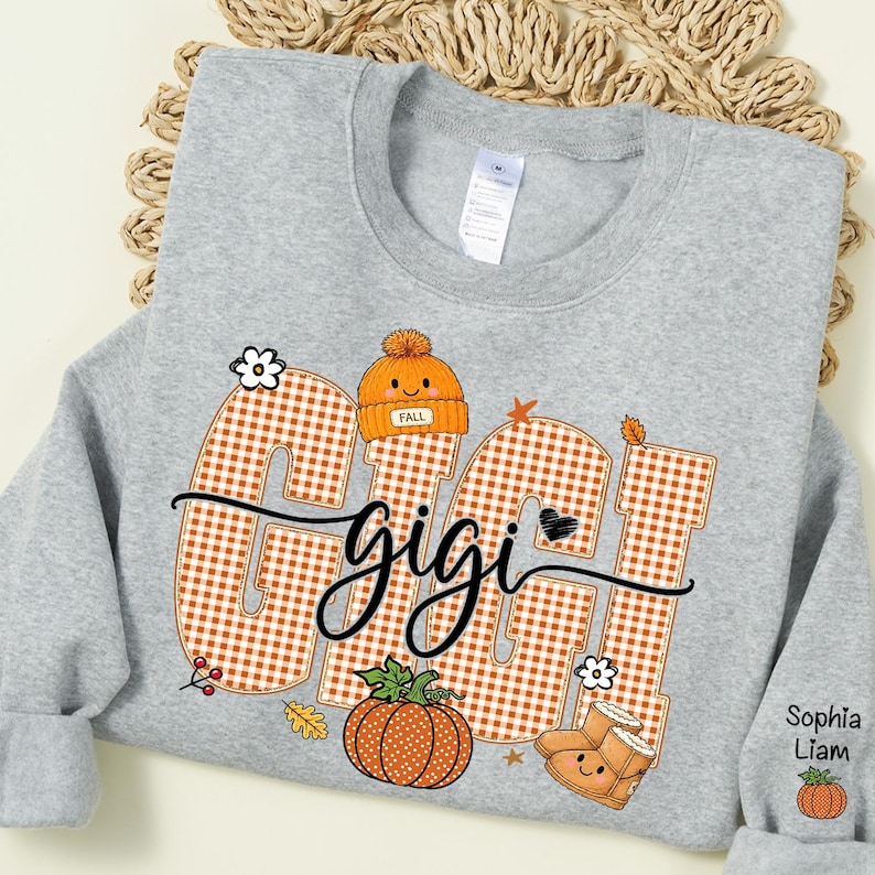 May include: A light gray crewneck sweatshirt featuring the word GIGI in large orange and white gingham letters. The design includes an orange beanie, a pumpkin, and boots with faces. The sleeve displays the names Sophia and Liam with a small pumpkin.
