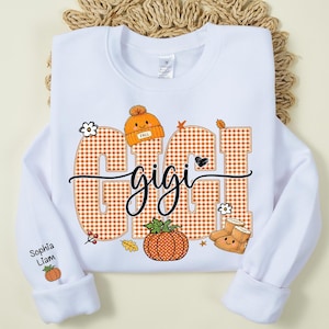 May include: A white crewneck sweatshirt featuring the text GIGI in large orange gingham letters. The design includes a cute orange beanie, a pumpkin, and tan boots with faces. The sleeve displays the names Sophia and Liam with a small pumpkin.