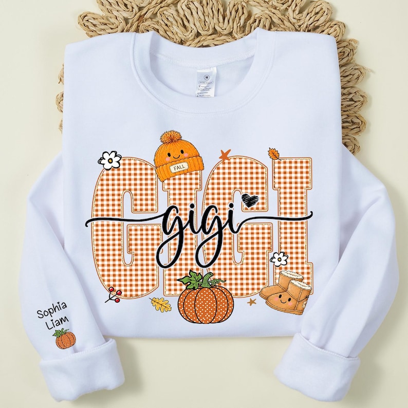 May include: A white crewneck sweatshirt featuring the text GIGI in large orange gingham letters. The design includes a cute orange beanie, a pumpkin, and tan boots with faces. The sleeve displays the names Sophia and Liam with a small pumpkin.