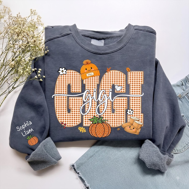 May include: A charcoal gray crewneck sweatshirt featuring a large orange and white gingham graphic that spells GIGI with a cursive overlay. The design includes a pumpkin, a beanie with the word FALL, and boots. The sleeve displays the names Sophia and Liam.