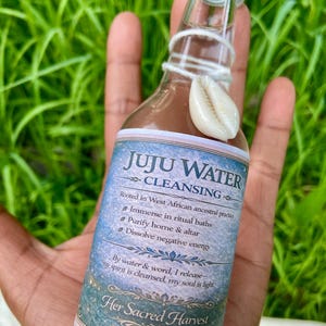 Peut inclure: Une bouteille en verre transparent avec un bouchon doré contenant un liquide, avec une étiquette indiquant JUJU WATER CLEANSING, Rooted in West African ancestral practice, Immerse in ritual baths, Purify home & altar, Dissolve negative energy, By water & word, I release—my spirit is cleansed, my soul is light, Her Sacred Harvest. Un coquillage cauri blanc est attaché au goulot avec une ficelle blanche.