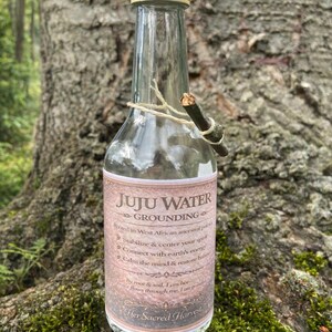 Può includere: Una bottiglia in vetro trasparente con tappo a vite chiaro, dotata di un'etichetta con la scritta JUJU WATER GROUNDING. L'etichetta include testo sui percorsi ancestrali dell'Africa occidentale e un piccolo rametto legato al collo con dello spago.