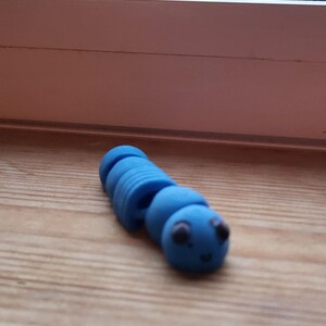 Può includere: Un giocattolo a forma di bruco blu con un corpo segmentato e un volto sorridente. Il giocattolo è realizzato in materiale morbido ed è lungo circa 7.6 cm. È appoggiato su una superficie di legno.